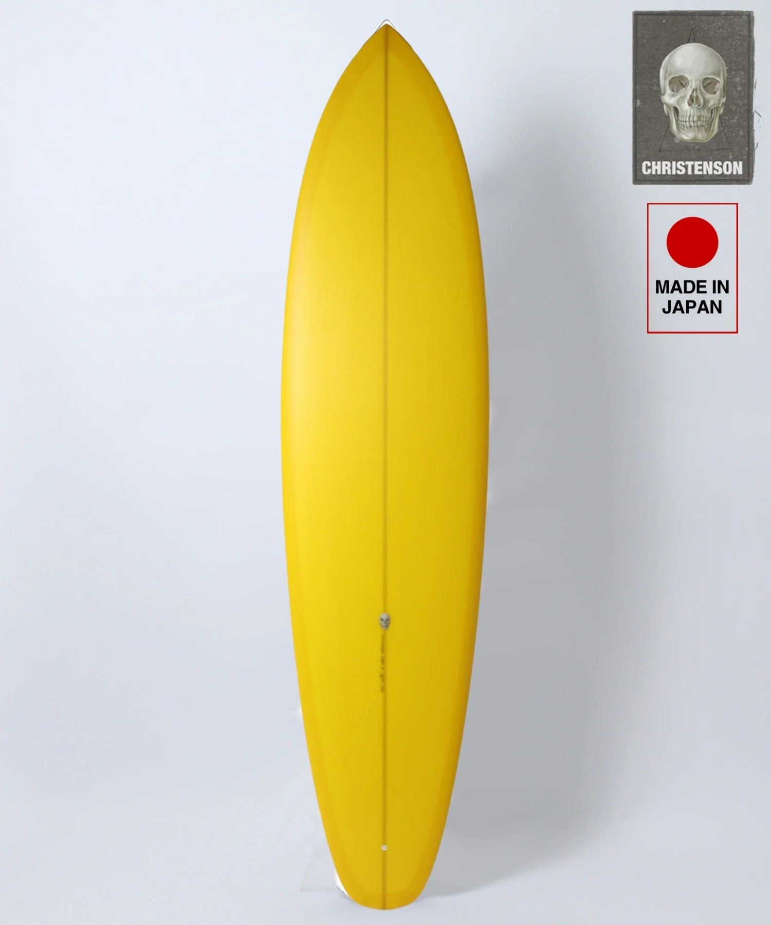 Yellow surfboard standing upright against a white background with two branding stickers on the right (Christenson skull card and Made in Japan label).