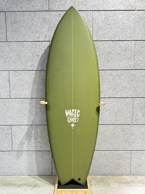 Olive-green surfboard standing upright on a wooden stand against a gray brick wall; white 'Magic' logo in the middle.