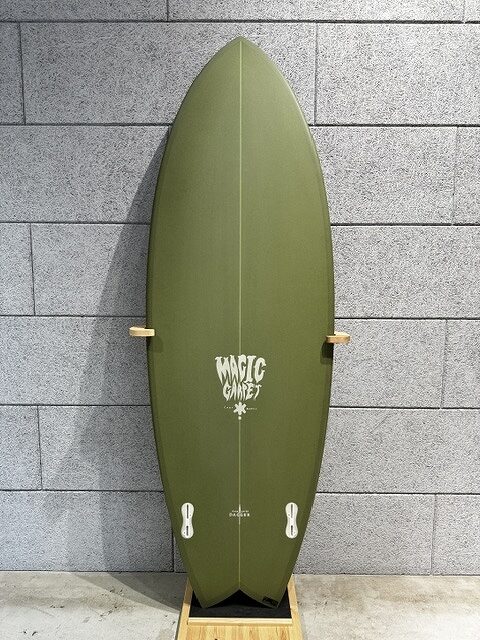 Olive-green surfboard with a pointed nose, standing upright on a wooden rack against a gray tiled wall, logo reads 'Magic Carvers' in the center.