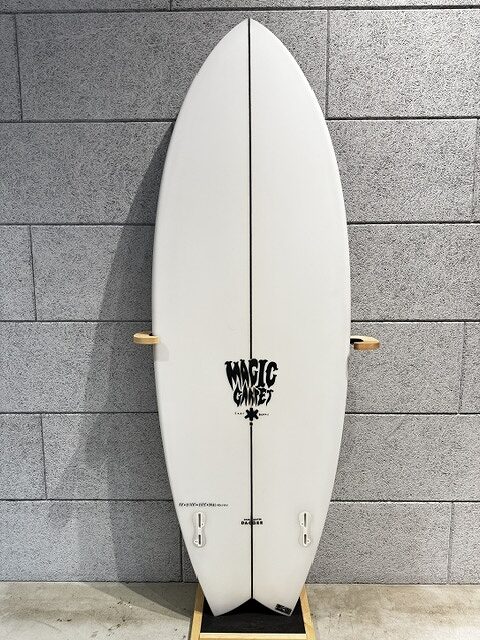 White surfboard with a black center stringer, resting on a wooden stand against a gray brick wall; displays the 'Magic Carpet' logo near the lower half.