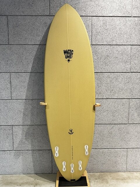Yellow surfboard standing upright on a wooden rack against a gray tiled wall, with a small black logo near the top.