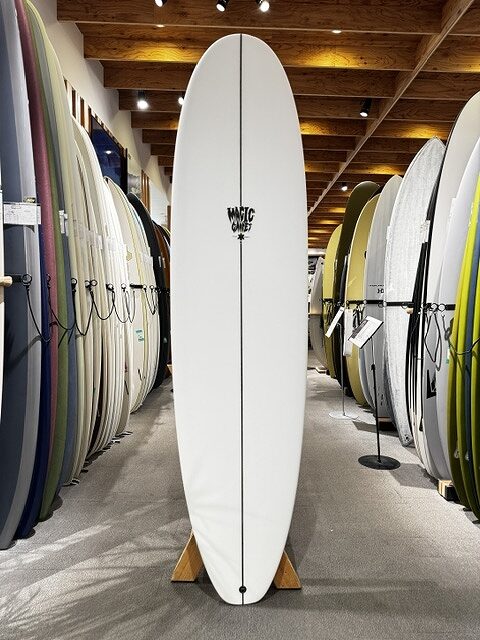 White surfboard standing upright on a display rack in a surf shop, with rows of colorful boards lining both sides of the aisle.