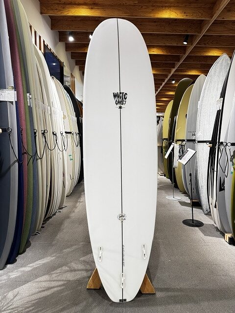 White surfboard standing upright on a display rack in a surf shop, surrounded by colorful boards under a wooden ceiling.