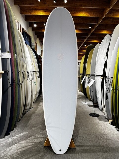 White surfboard standing upright on a wooden stand in a surf shop, surrounded by rows of other boards.