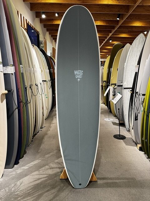 Gray surfboard on a wooden stand in a shop, surrounded by rows of colorful surfboards on racks.