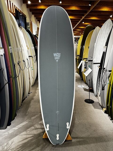 Center-standing gray surfboard on a stand in a shop, surrounded by rows of other boards along the walls.