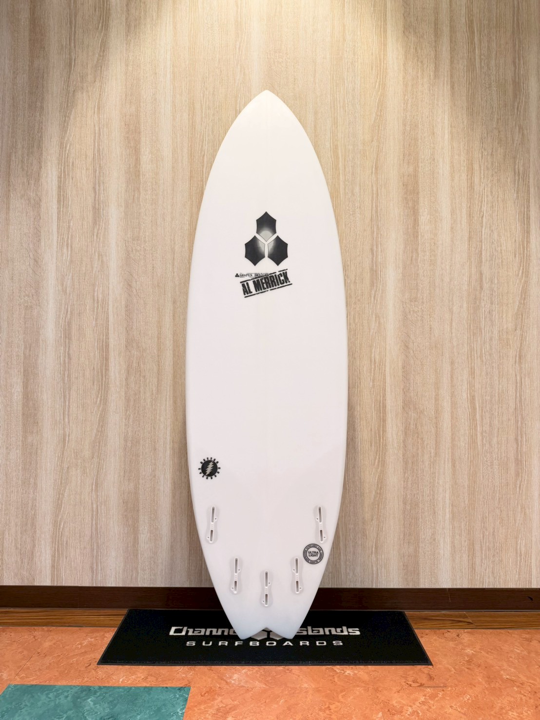 White surfboard on a display stand against a wood-paneled wall, showing multiple fin boxes and logos (Al Merrick)