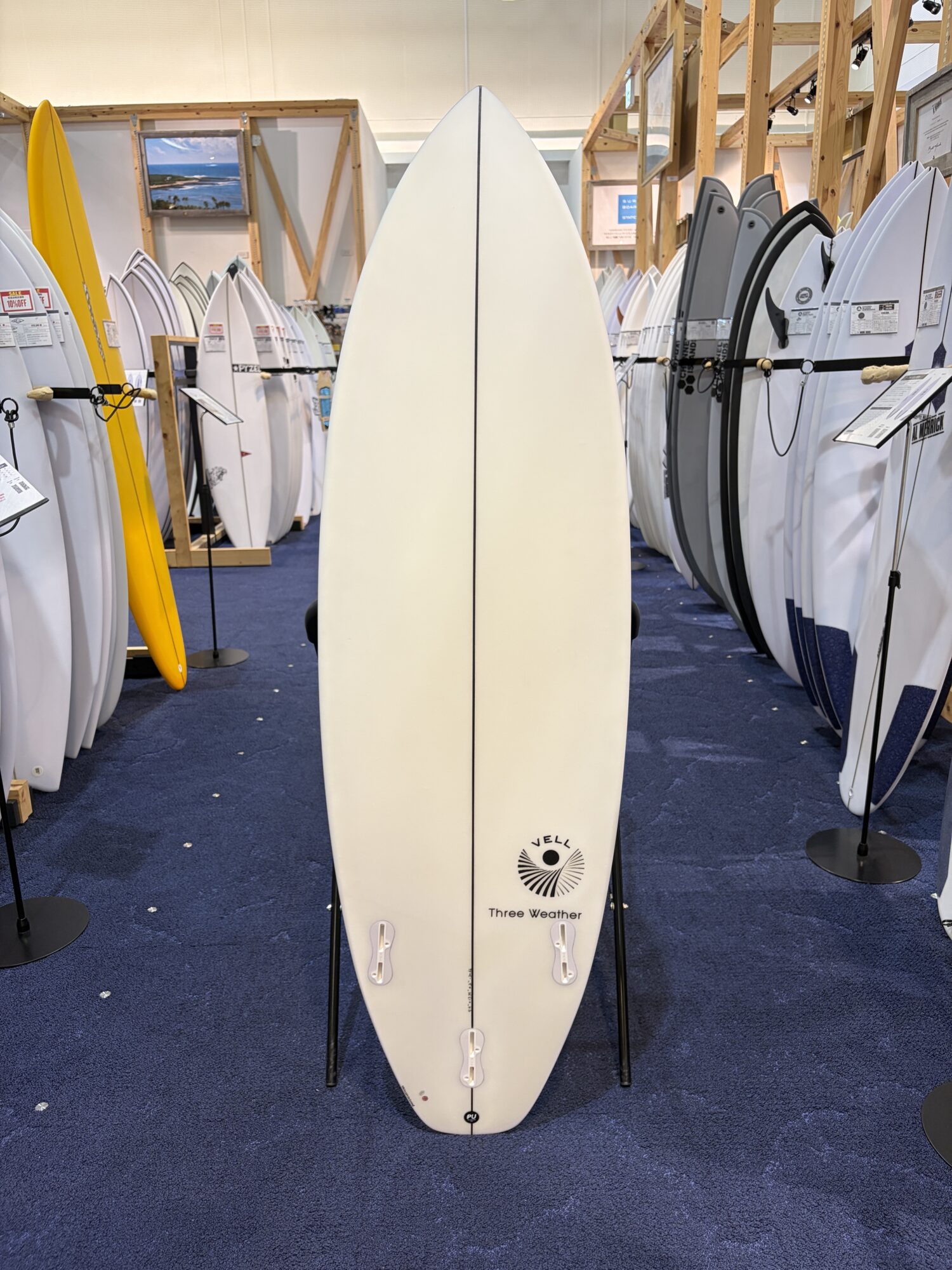 IMG_6166 - SHOP NEWS | ムラサキスポーツオンラインストア 通販 White surfboard standing upright in a crowded showroom with many boards on display around it.