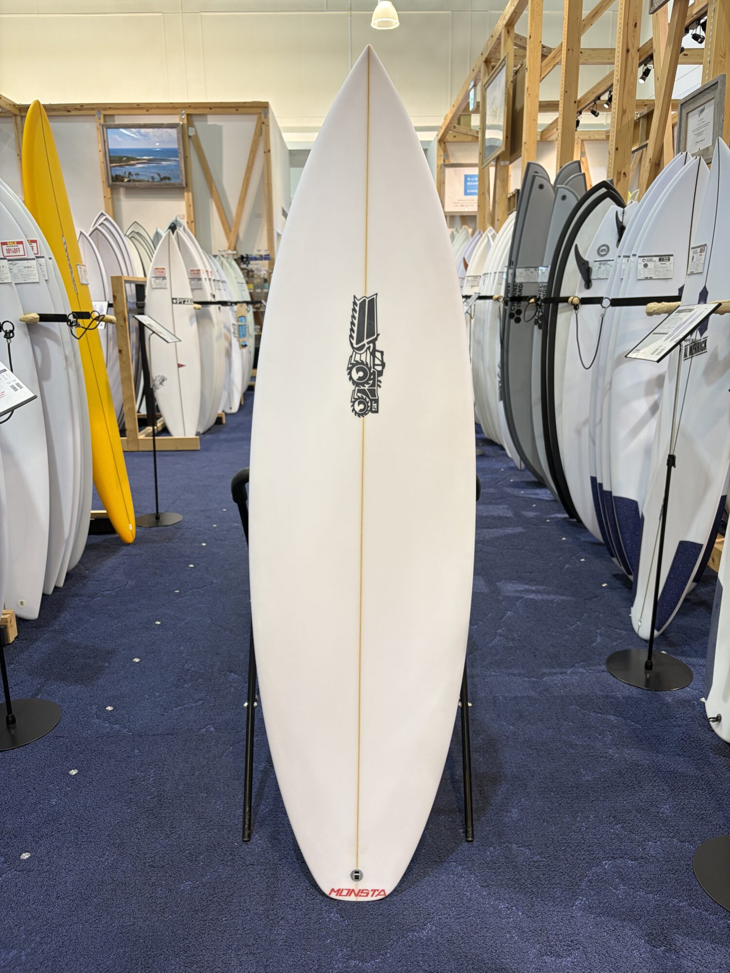 IMG_6167 - SHOP NEWS | ムラサキスポーツオンラインストア 通販 White surfboard with a Monsta logo stands upright on two supports in a showroom filled with surfboards on racks nearby.