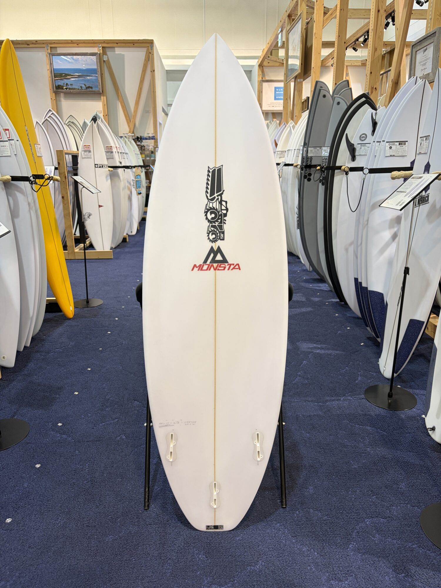 IMG_6168 - SHOP NEWS | ムラサキスポーツオンラインストア 通販 White surfboard on a stand in a showroom, MONSTA logo visible near the bottom center, surrounded by many other boards on racks.