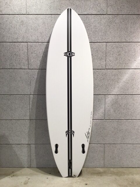 s-image3 - SHOP NEWS | ムラサキスポーツオンラインストア 通販 White surfboard with a black center stripe, propped upright against a gray tiled wall; logos along the center and near the tail, fin boxes visible.
