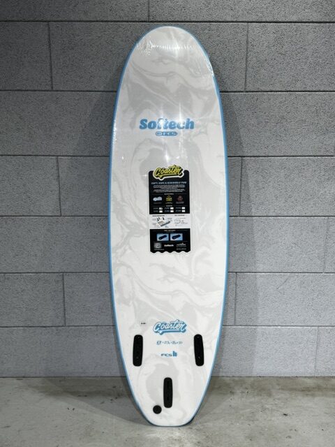 White surfboard with blue edge trim leaning against a gray tiled wall, displaying brand stickers and fin boxes at the bottom.