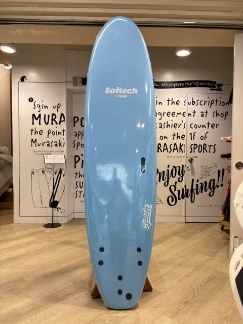Blue Softtech surfboard on a wooden stand, displayed indoors with shop signage in the background.