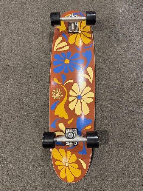 Top-down view of a longboard with an orange deck covered in yellow and blue hibiscus flowers and black wheels.",