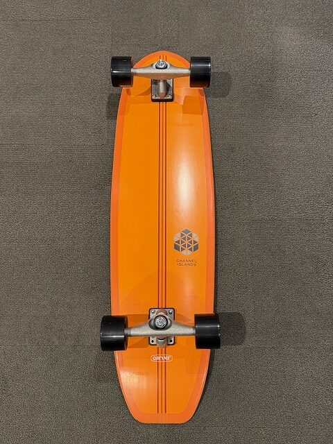 Top-down view of an orange longboard with black wheels and silver trucks on a dark carpet.