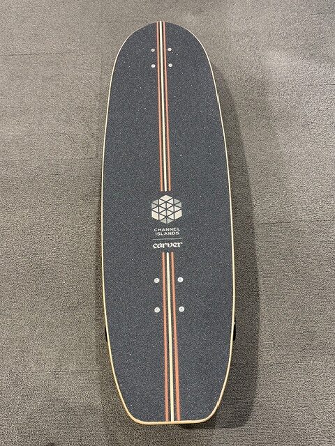 Longboard skateboard deck on a dark floor with red and cream stripes down the center and a geometric logo in the middle read as Channel Islands Carver? text (board graphic)