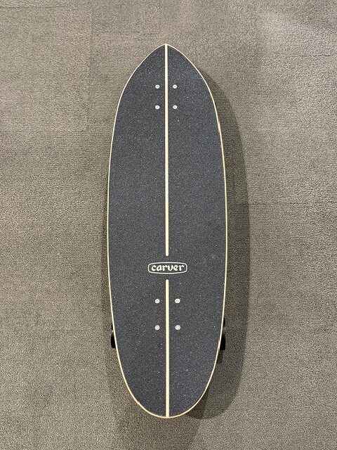 Top-down view of a Carver longboard skateboard with a dark deck, pale edge trim, and a central light stripe featuring multiple mounting holes near the nose and tail.