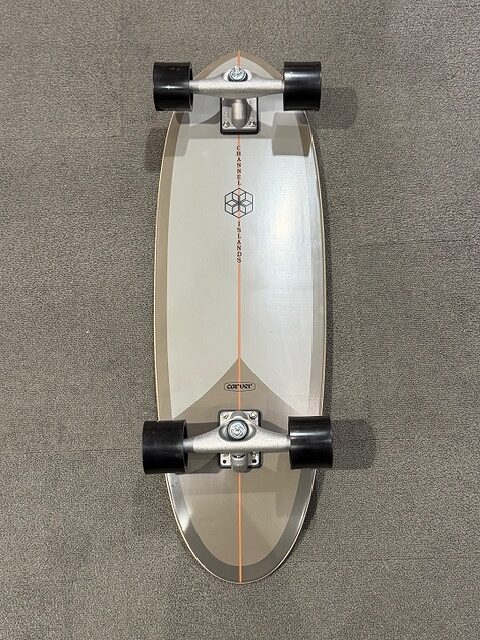 Top-down view of a gray longboard with an orange center line and black wheels on a gray carpeted floor.