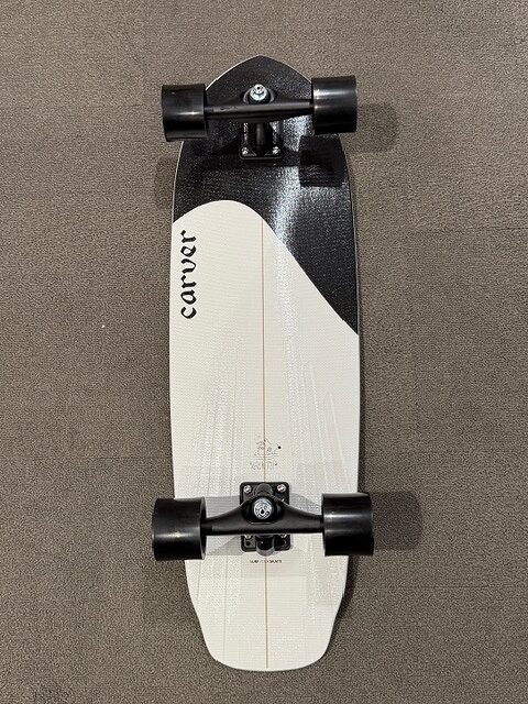 Top-down view of a Carver longboard with a white deck and black trucks and wheels on a grey surface.
