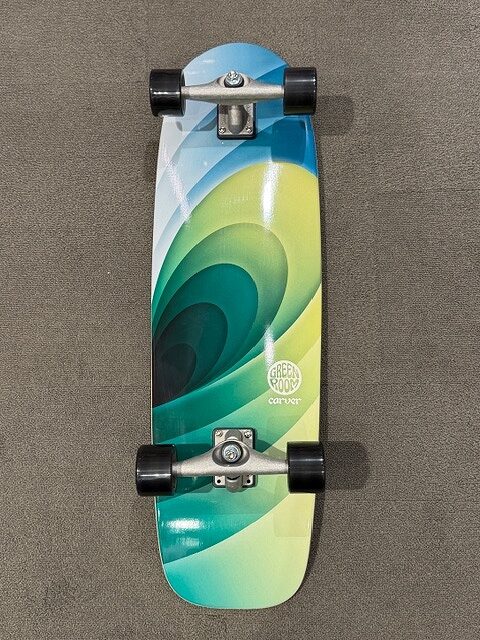 Longboard skateboard with a blue–green–yellow swirl graphic, four black wheels and silver trucks on a carpeted surface.
