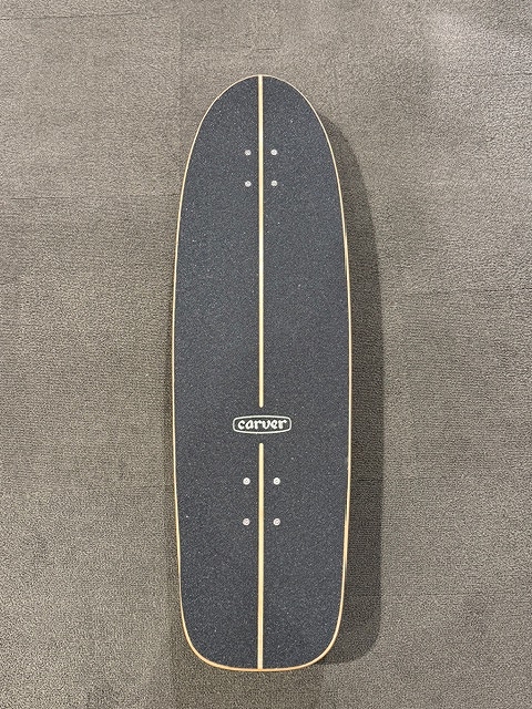 Vertical view of a dark longboard/deck with light wooden rails and a centered line, standing on a carpeted floor in a minimalist pose.