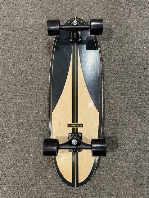 Top-down view of a Carver longboard with black trucks and five-spoke wheels on a carpeted floor.
