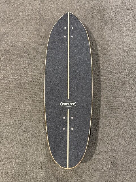 Top-down view of a Carver longboard deck with dark blue grip tape, natural wood rails, and a central beige stripe across the length.