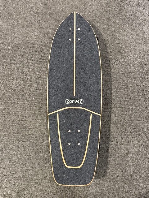 Top-down view of a black Carver longboard with beige rails, lying on a gray carpeted surface.