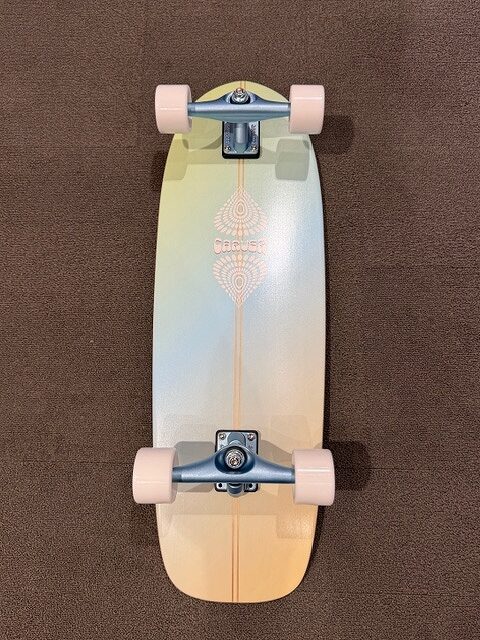 Top-down view of a pastel gradient longboard with white wheels and silver trucks on a brown carpet.
