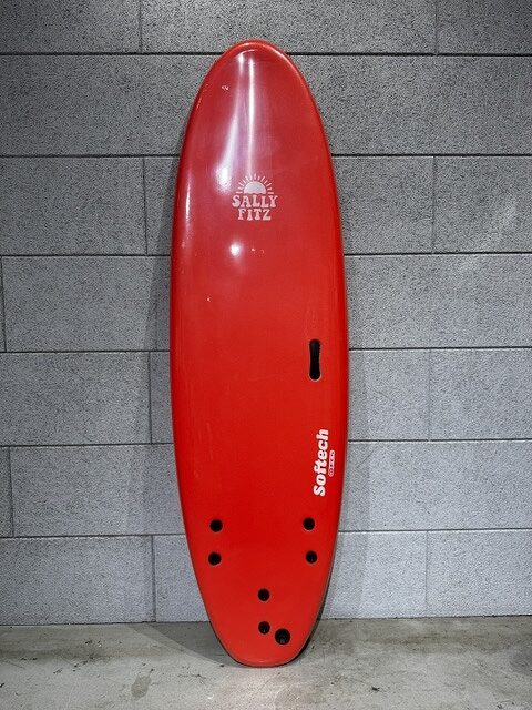 Red shortboard surfboard leaning upright against a gray brick wall, with visible fin boxes and multiple fin holes near the tail, branded 'SALLY FITZ' and 'Softech'.