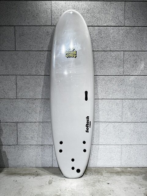 White surfboard standing upright against a gray tiled wall, with several tail holes and a fin slot; branding decals visible on the board.