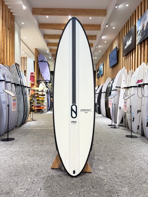 White surfboard with a black center stripe and Mindcraft logo, displayed upright in a surf shop aisle.
