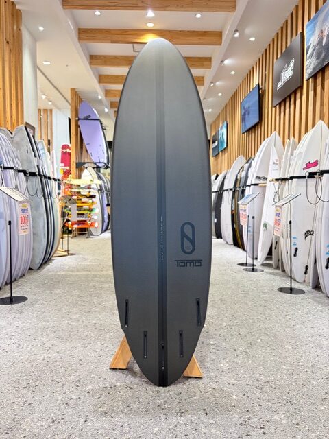 Tall gray surfboard on a display stand in a surf shop showroom, with numerous boards lined along the walls and wooden beams overhead.