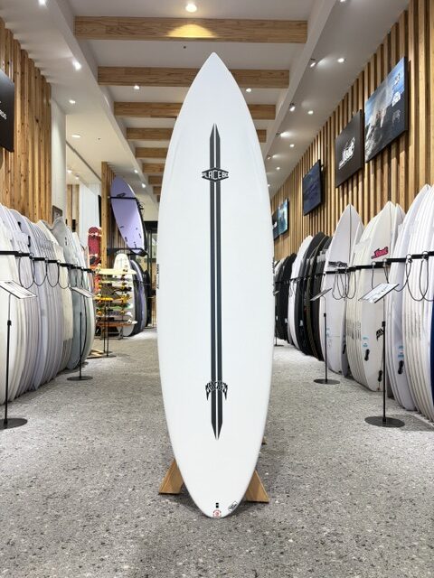 Tall white surfboard on a display rack in a surf shop, centered between rows of other boards, with a vertical stripe and logo down the center.