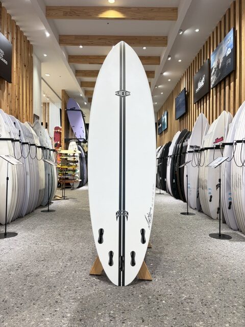 White surfboard with two black stripes standing upright on a wooden rack inside a surf shop showroom, surrounded by rows of other boards on racks.