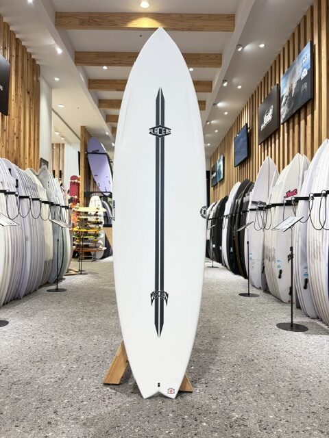 Tall white surfboard with a vertical black stripe and logos stands upright on a wooden rack in a surf shop, surrounded by rows of other surfboards.