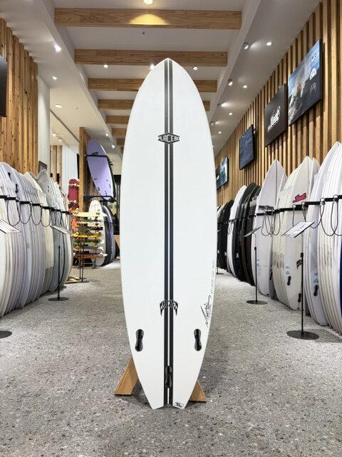 Tall white surfboard with black rails and logo, standing upright on a wooden stand inside a surf shop showroom surrounded by boards on both sides.