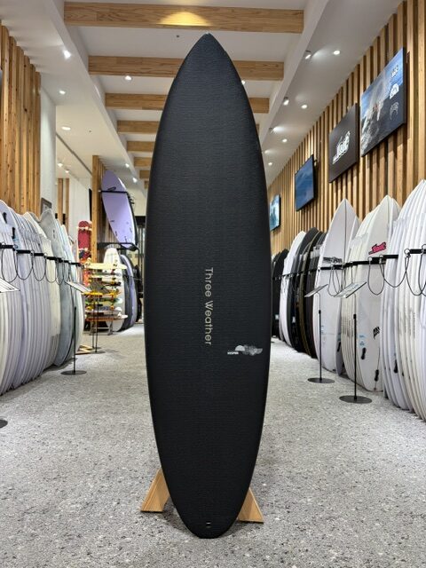 Tall black surfboard standing upright on a wooden stand in a surf shop, with white boards lining both sides of the aisle.