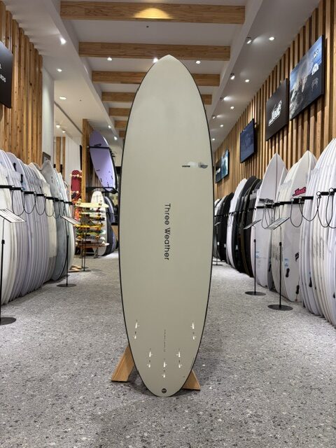 Cream-colored surfboard standing upright on a wooden stand in a surf shop showroom, with many boards lined up along both walls on racks.