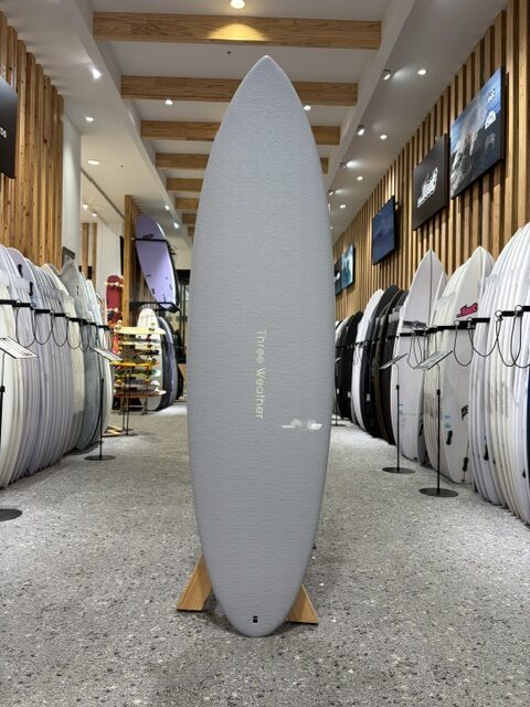 Gray surfboard standing upright on a small wooden stand in a surf shop aisle, with rows of surfboards on racks along both walls and wall-mounted displays in the background.