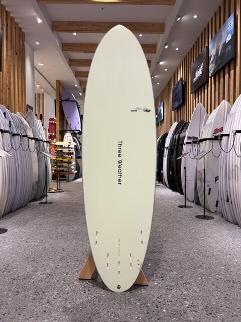 Cream-colored surfboard on a wooden stand in a surf shop, surrounded by rows of other boards on racks.