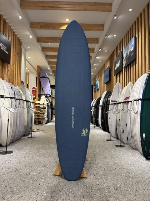 Blue longboard surfboard on a wooden stand in a surf shop, surrounded by rows of boards on racks.