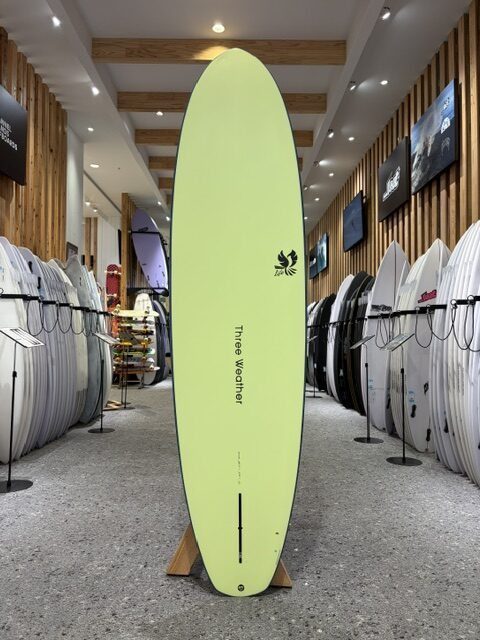 Bright lime-green surfboard on a wooden stand in a showroom, with 'Tree Weather' text down the center, surrounded by rows of other boards.