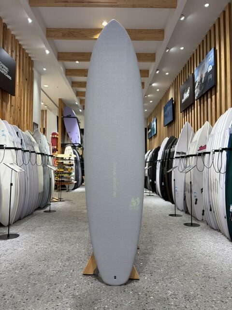 Tall gray surfboard standing upright on a wooden stand in a bright showroom, with racks of boards lining both sides.