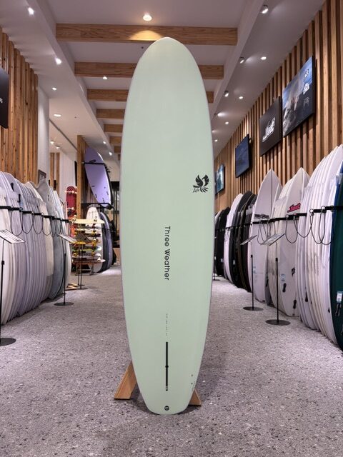 Mint-green surfboard on a wooden stand in a surf shop, surrounded by racks of white and colored boards along wood-paneled walls.