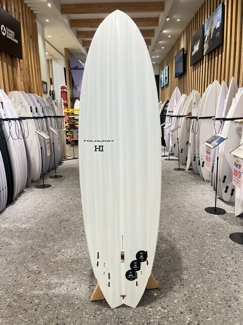 Tall white surfboard on a display stand in a showroom, surrounded by rows of other surfboards against wooden walls.