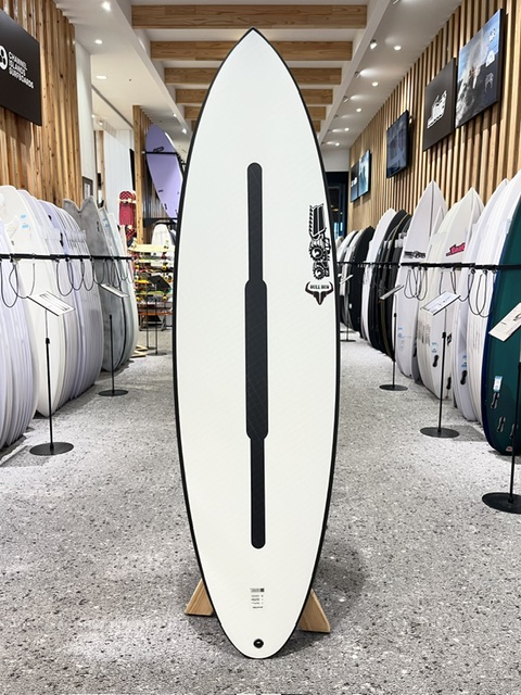 White surfboard on a stand inside a surf shop, with racks of boards lining both sides behind it.