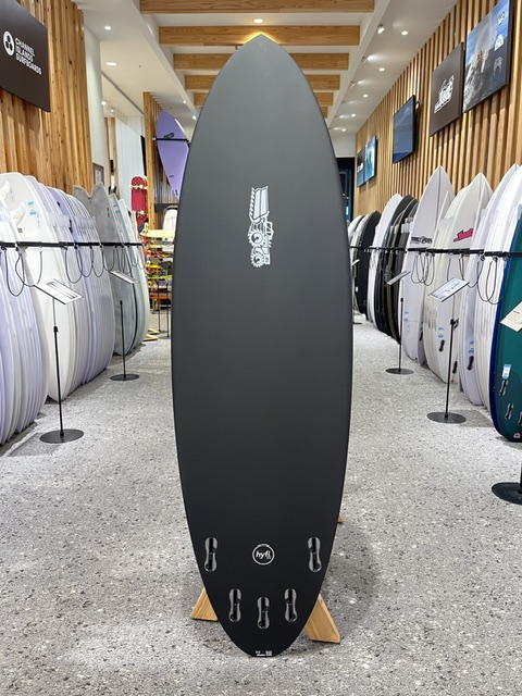 Dark gray surfboard standing upright on a display rack in a surf shop, surrounded by other boards on both sides.