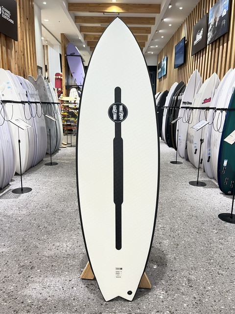 White surfboard with a black vertical stripe and logo standing upright on a display rack in a surf shop.