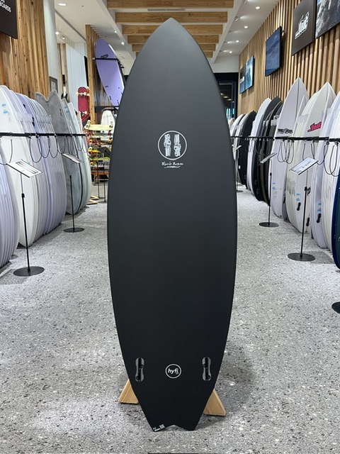 Matte black surfboard on a wooden stand in a surf shop, surrounded by other boards and wooden accents.